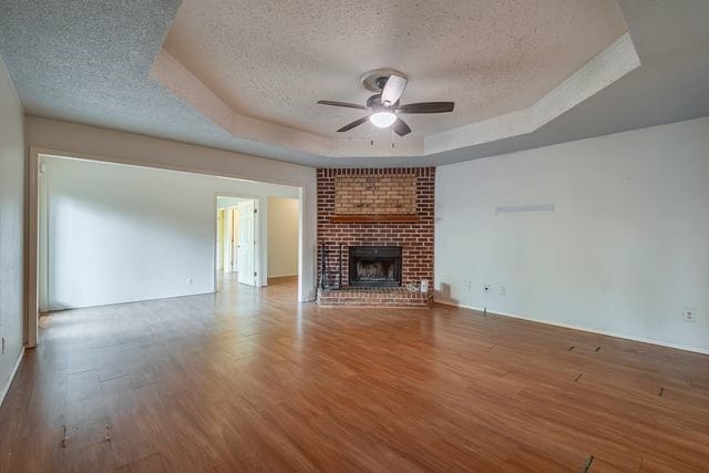 unfurnished living room featuring a tray ceiling, a fireplace, a ceiling fan, a textured ceiling, and wood finished floors