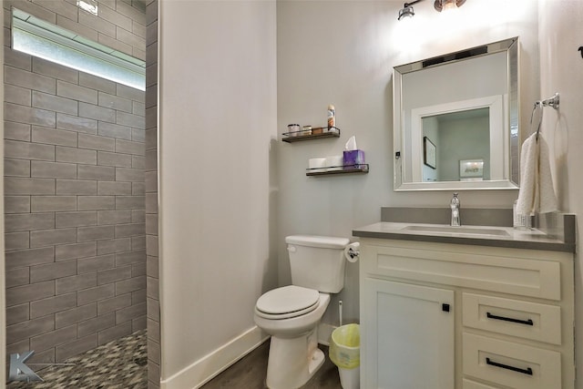 bathroom with toilet, wood finished floors, vanity, baseboards, and tiled shower