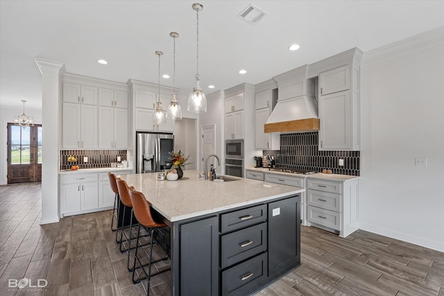 kitchen with custom exhaust hood, white cabinetry, stainless steel appliances, wood-type flooring, and an island with sink