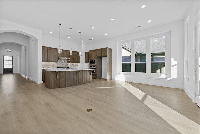 kitchen with appliances with stainless steel finishes, hanging light fixtures, light wood-type flooring, backsplash, and an island with sink