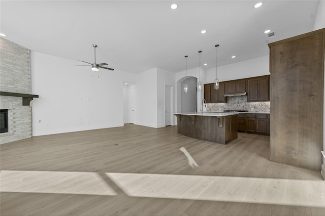 kitchen featuring a fireplace, tasteful backsplash, hanging light fixtures, a kitchen island with sink, and light wood-type flooring