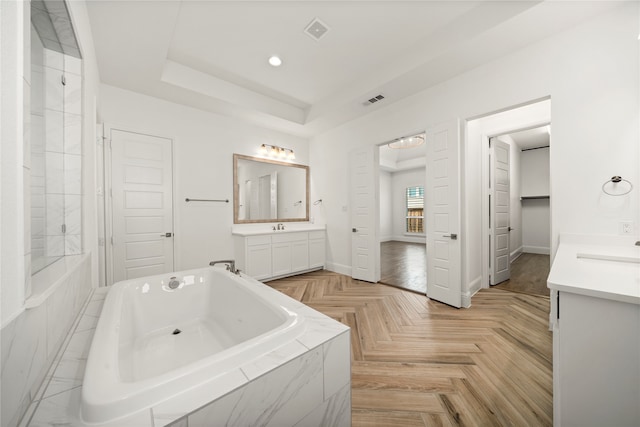 bathroom with a tray ceiling, vanity, parquet floors, and a relaxing tiled tub