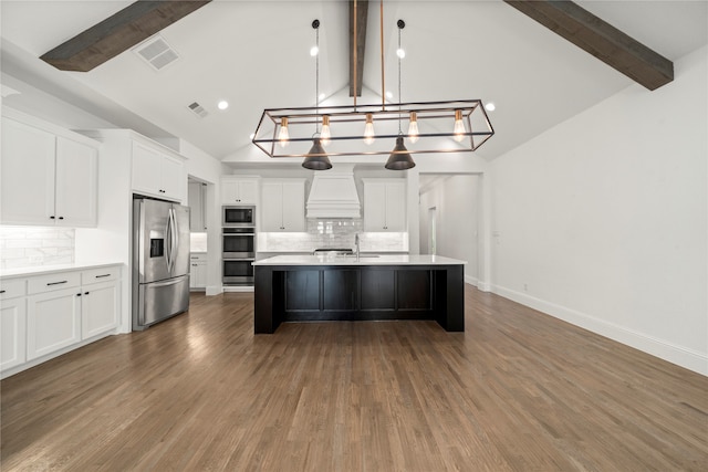 kitchen featuring beamed ceiling, premium range hood, an island with sink, pendant lighting, and appliances with stainless steel finishes