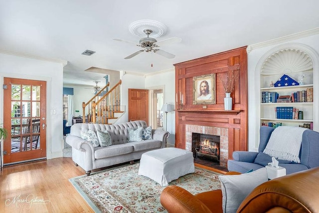 living room featuring ceiling fan, built in shelves, a brick fireplace, ornamental molding, and light hardwood / wood-style floors