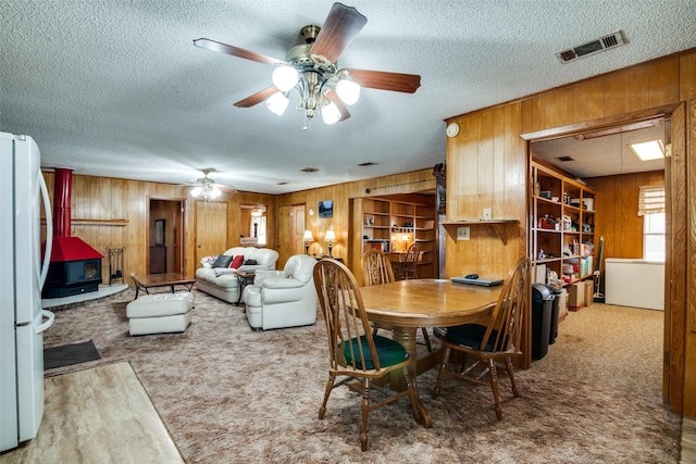 carpeted dining space with ceiling fan, wooden walls, a textured ceiling, and a wood stove