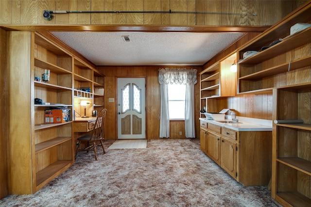 kitchen with a textured ceiling, built in desk, sink, wood walls, and light colored carpet