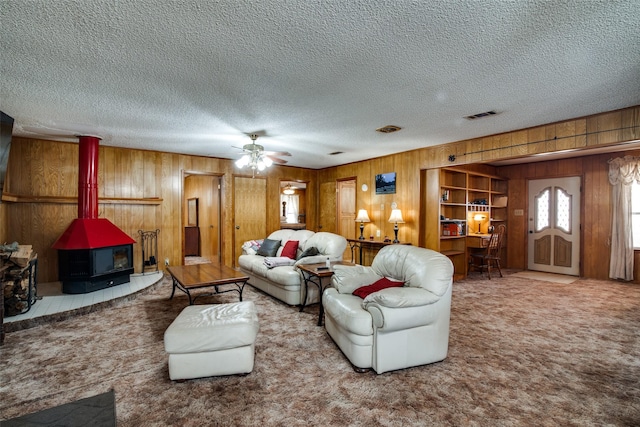 living room with a textured ceiling, carpet floors, wooden walls, a wood stove, and ceiling fan