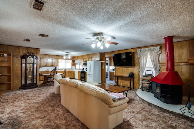 living room featuring ceiling fan, light colored carpet, a wood stove, a textured ceiling, and wooden walls