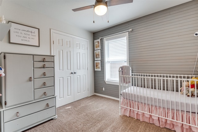 carpeted bedroom featuring a crib, wood walls, ceiling fan, and a closet