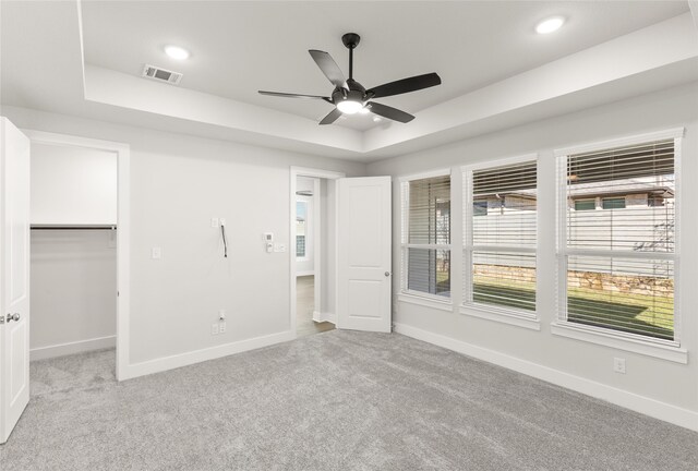 unfurnished bedroom with light carpet, a walk in closet, a raised ceiling, ceiling fan, and a closet