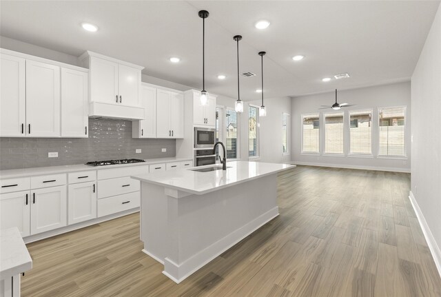 kitchen featuring appliances with stainless steel finishes, light wood-type flooring, white cabinetry, decorative light fixtures, and a center island with sink