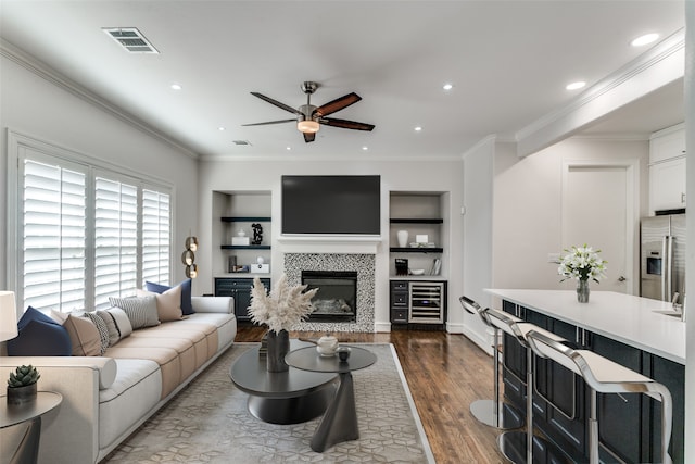 living room featuring a tiled fireplace, ceiling fan, dark hardwood / wood-style floors, built in shelves, and crown molding