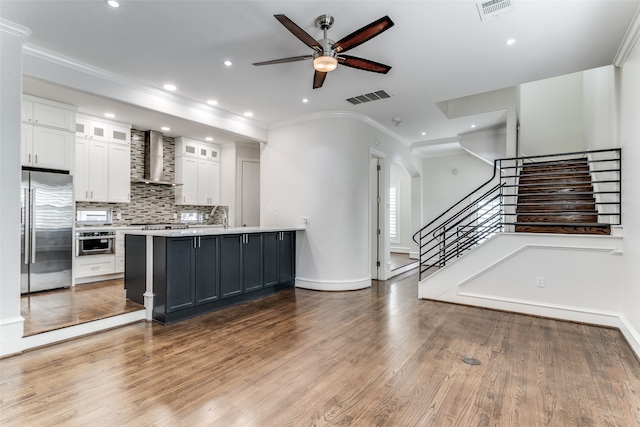 kitchen with a large island with sink, wall chimney range hood, wood-type flooring, white cabinetry, and appliances with stainless steel finishes