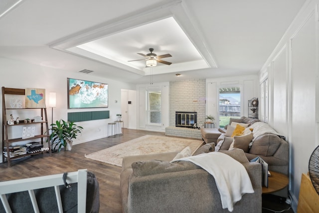 living room with brick wall, a raised ceiling, dark hardwood / wood-style flooring, ceiling fan, and a brick fireplace