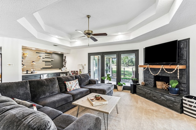 living room with a textured ceiling, a stone fireplace, light carpet, a tray ceiling, and ceiling fan