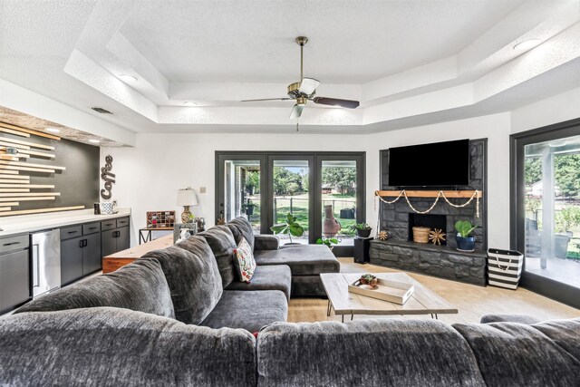 living room with a textured ceiling, ceiling fan, a stone fireplace, and a tray ceiling