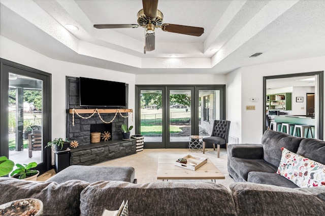 living room featuring ceiling fan, a stone fireplace, carpet floors, and a tray ceiling