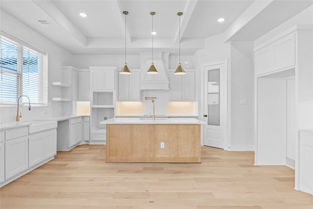 kitchen with light wood-type flooring, an island with sink, hanging light fixtures, white cabinetry, and custom range hood