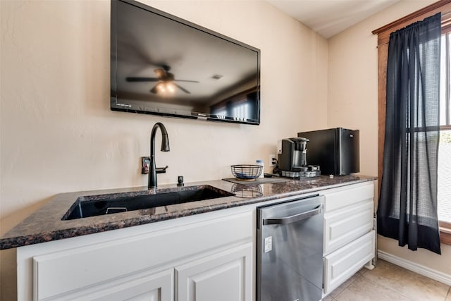 kitchen with light tile patterned floors, stainless steel dishwasher, white cabinets, a sink, and dark stone counters