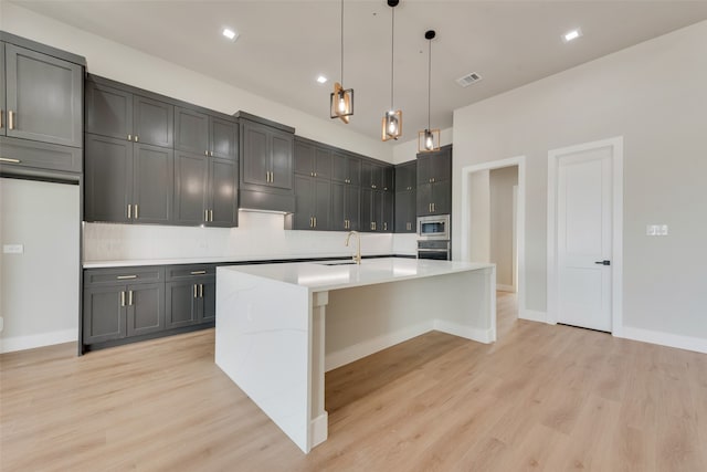 kitchen with decorative light fixtures, appliances with stainless steel finishes, light hardwood / wood-style flooring, decorative backsplash, and a center island with sink