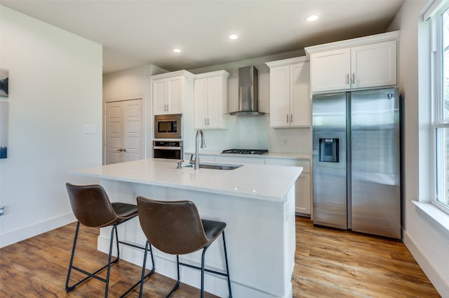 kitchen with wall chimney exhaust hood, a kitchen island with sink, light hardwood / wood-style floors, stainless steel appliances, and sink