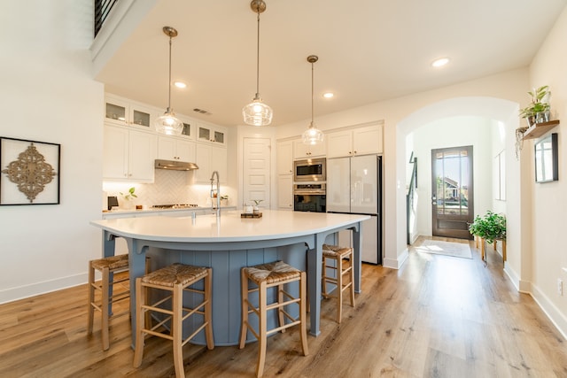 kitchen with appliances with stainless steel finishes, decorative light fixtures, light hardwood / wood-style flooring, white cabinets, and an island with sink