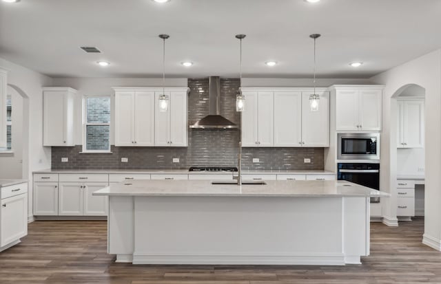 kitchen featuring appliances with stainless steel finishes, wall chimney exhaust hood, decorative light fixtures, white cabinetry, and an island with sink