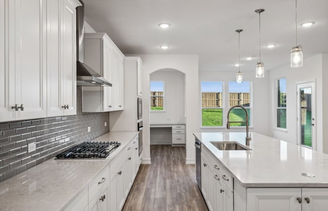 kitchen with sink, wall chimney exhaust hood, decorative light fixtures, a kitchen island with sink, and appliances with stainless steel finishes