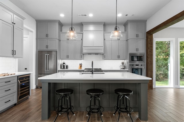 kitchen with pendant lighting, wine cooler, an island with sink, gray cabinets, and appliances with stainless steel finishes