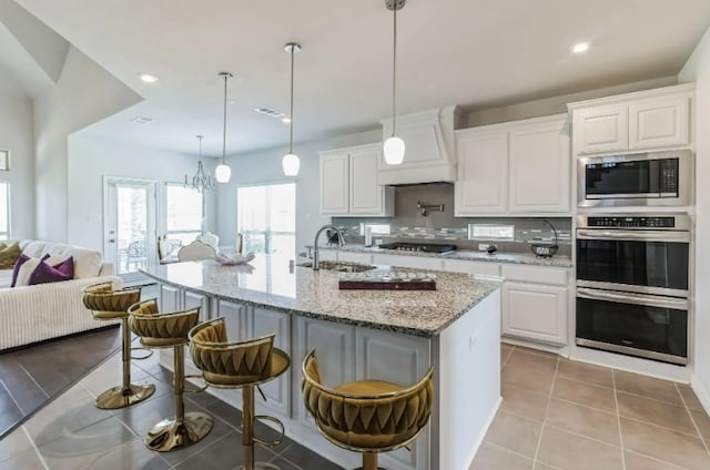 kitchen with white cabinets, stainless steel appliances, light stone counters, an island with sink, and light tile patterned flooring