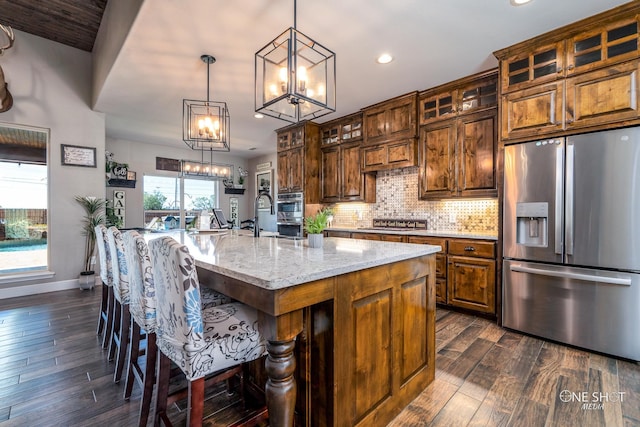 kitchen with a kitchen island with sink, hanging light fixtures, light stone counters, appliances with stainless steel finishes, and dark hardwood / wood-style flooring