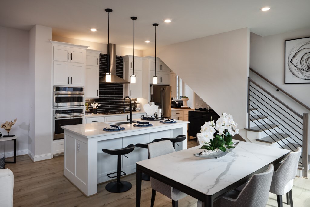 kitchen with wall chimney range hood, light wood-type flooring, an island with sink, stainless steel appliances, and pendant lighting