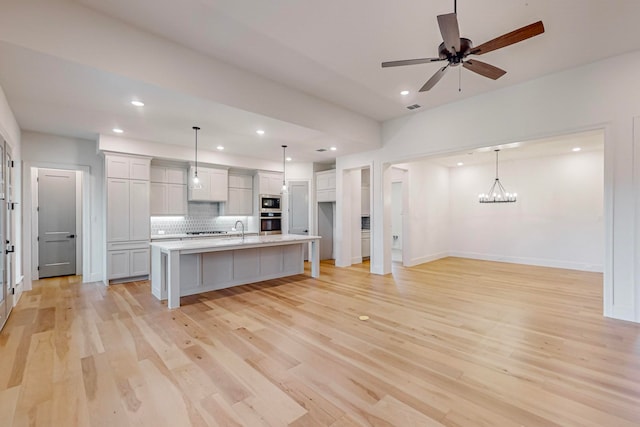 kitchen featuring light hardwood / wood-style floors, ceiling fan with notable chandelier, backsplash, decorative light fixtures, and a center island with sink