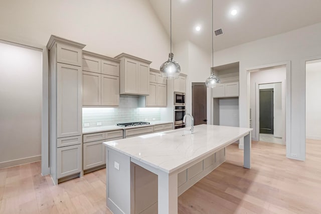 kitchen with light stone counters, hanging light fixtures, a kitchen island with sink, light hardwood / wood-style flooring, and appliances with stainless steel finishes