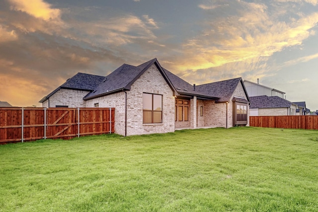 back of property at dusk featuring a fenced backyard, a yard, brick siding, and roof with shingles