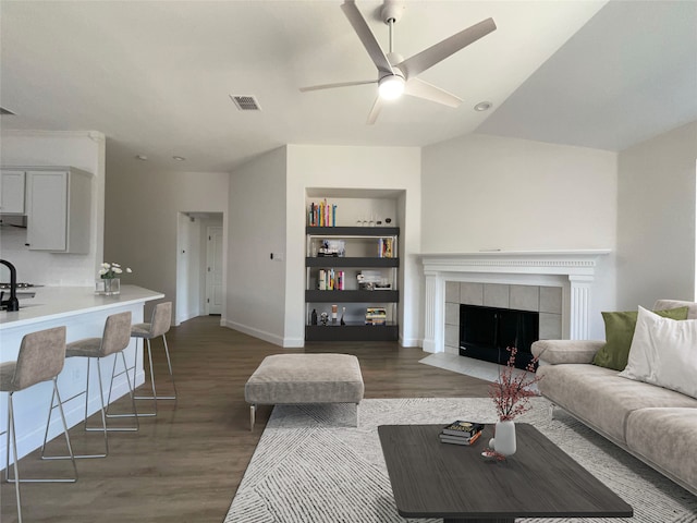 living room with a tiled fireplace, vaulted ceiling, built in shelves, dark wood-type flooring, and ceiling fan