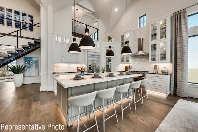 kitchen with high vaulted ceiling, a center island with sink, wall chimney range hood, decorative light fixtures, and white cabinetry