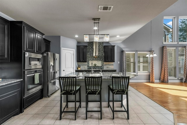 kitchen with a center island with sink, hanging light fixtures, light stone countertops, appliances with stainless steel finishes, and light hardwood / wood-style floors