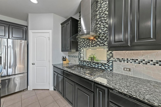 kitchen with backsplash, wall chimney range hood, dark stone countertops, light tile patterned floors, and appliances with stainless steel finishes