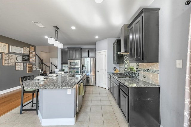kitchen with wall chimney range hood, appliances with stainless steel finishes, dark stone counters, decorative backsplash, and light tile patterned floors