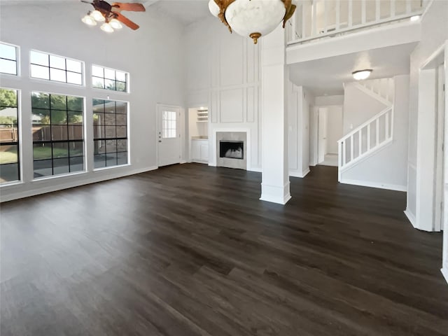 unfurnished living room featuring dark wood finished floors, a fireplace, a ceiling fan, baseboards, and stairs