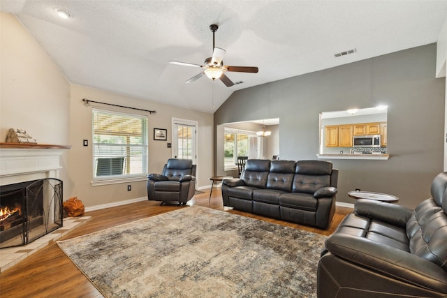 living area featuring a warm lit fireplace, visible vents, ceiling fan, wood finished floors, and vaulted ceiling