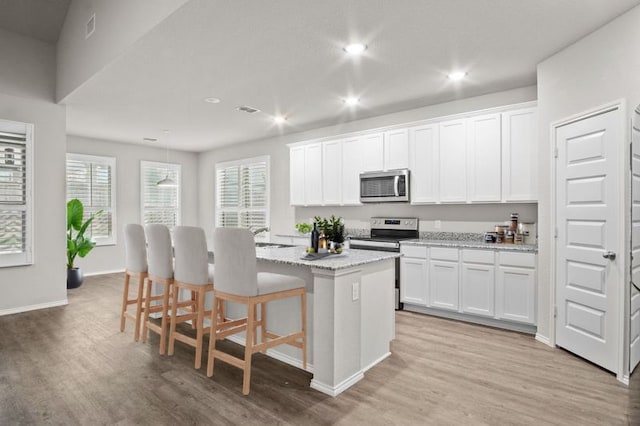 kitchen with light stone countertops, light hardwood / wood-style flooring, an island with sink, stainless steel appliances, and white cabinetry