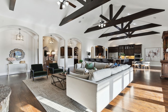 living room featuring high vaulted ceiling, arched walkways, dark wood-style flooring, beamed ceiling, and ceiling fan with notable chandelier