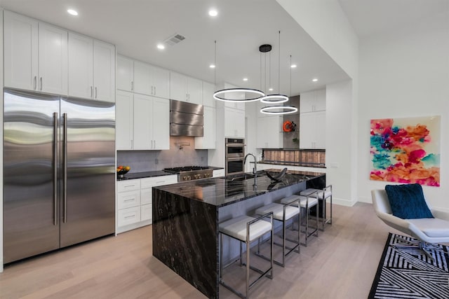 kitchen featuring appliances with stainless steel finishes, white cabinetry, a kitchen breakfast bar, a center island with sink, and dark stone counters