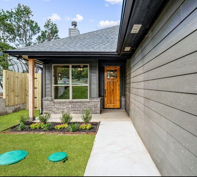 view of exterior entry with brick siding, fence, roof with shingles, a lawn, and a chimney