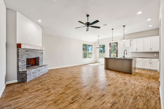 kitchen featuring a brick fireplace, light wood-type flooring, ceiling fan with notable chandelier, white cabinetry, and a kitchen island with sink