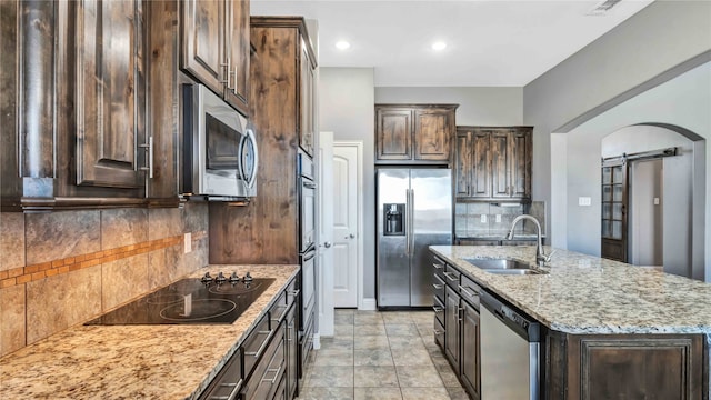 kitchen with light stone counters, a barn door, stainless steel appliances, a center island with sink, and sink