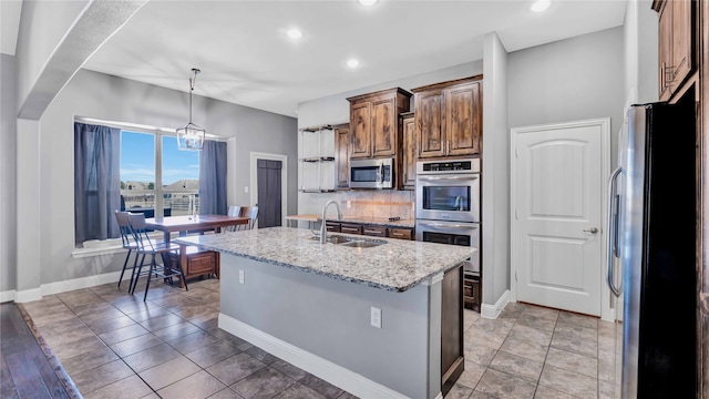 kitchen featuring a kitchen island with sink, sink, stainless steel appliances, decorative light fixtures, and light stone countertops