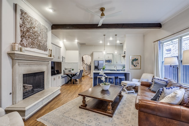 living room featuring light wood-type flooring, sink, beam ceiling, a premium fireplace, and ceiling fan